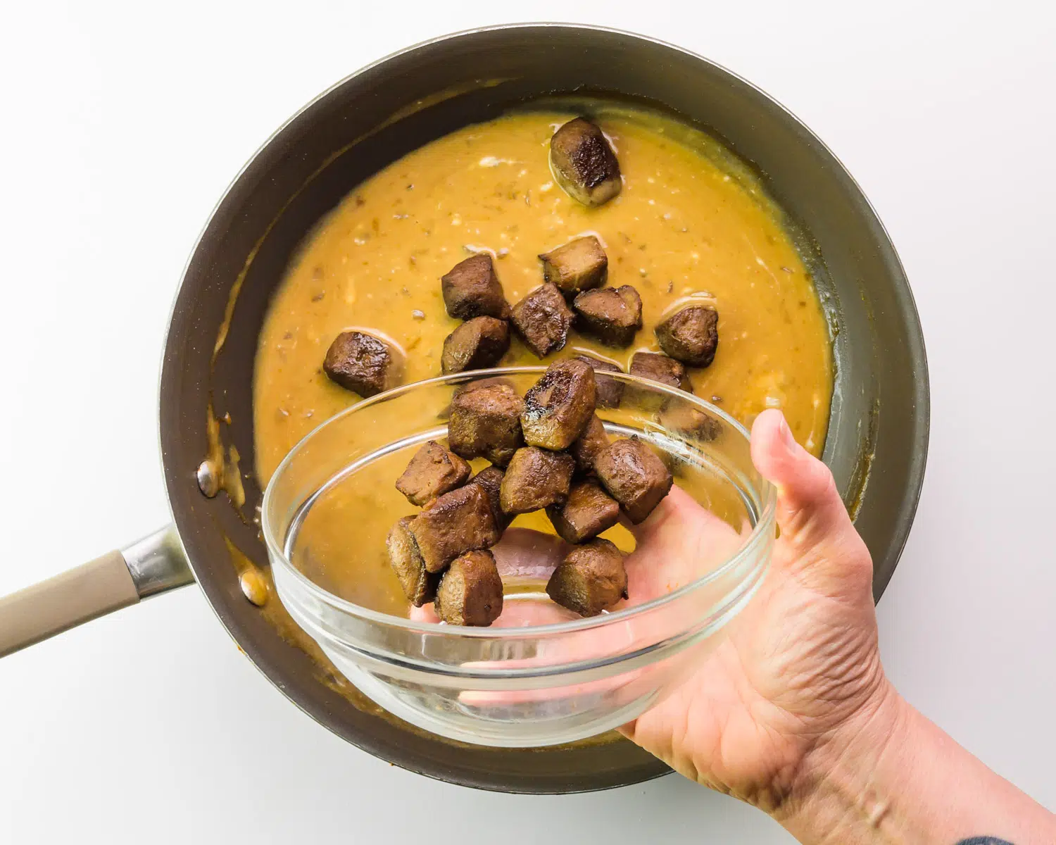 A hand holds a bowl of cooked vegan beef, adding them to a skillet with a creamy brown sauce.