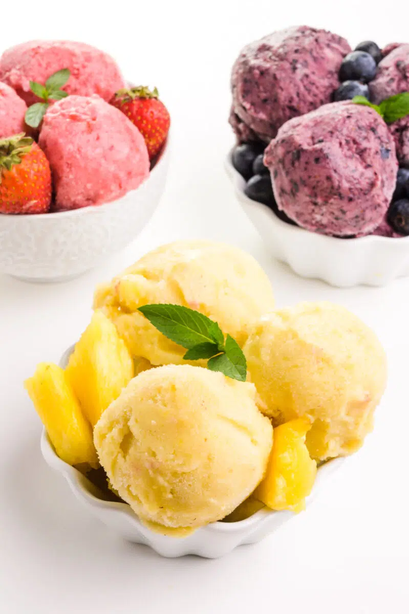 A bowl of pineapple frozen yogurt sits in front of bowls of blueberry and strawberry frozen yogurt.