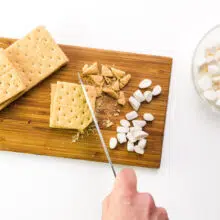 A hand holds a knife chopping graham crackers and marshmallows on a cutting board.