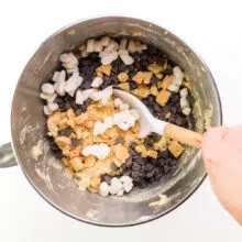 A hand stirs marshmallows and chocolate chips into cookie dough.
