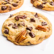 A chocolate chip pecan cookie sits on a white table. There are other cookies in the background.