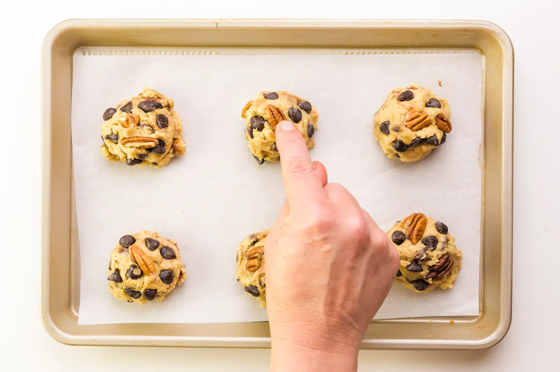 A hand presses a pecan on top of cookie dough balls on a cookie sheet.