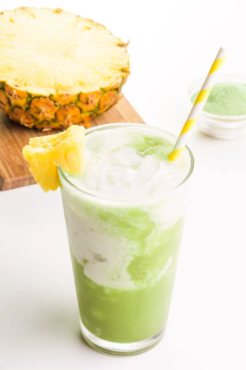 An iced pineapple matcha beverage has a yellow straw. There is green matcha powder and a sliced pineapple on a cutting board in the background.