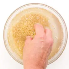 A hand reaches into a bowl with uncooked rice and water, stirring the rice.