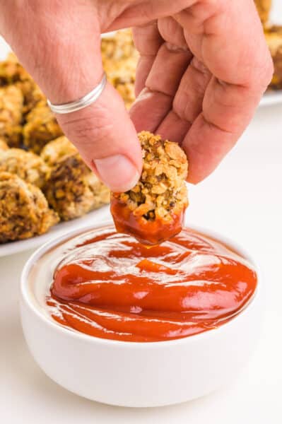 A hand holds a cauliflower tot after dipping it in a bowl of ketchup. There are more tots in the background.