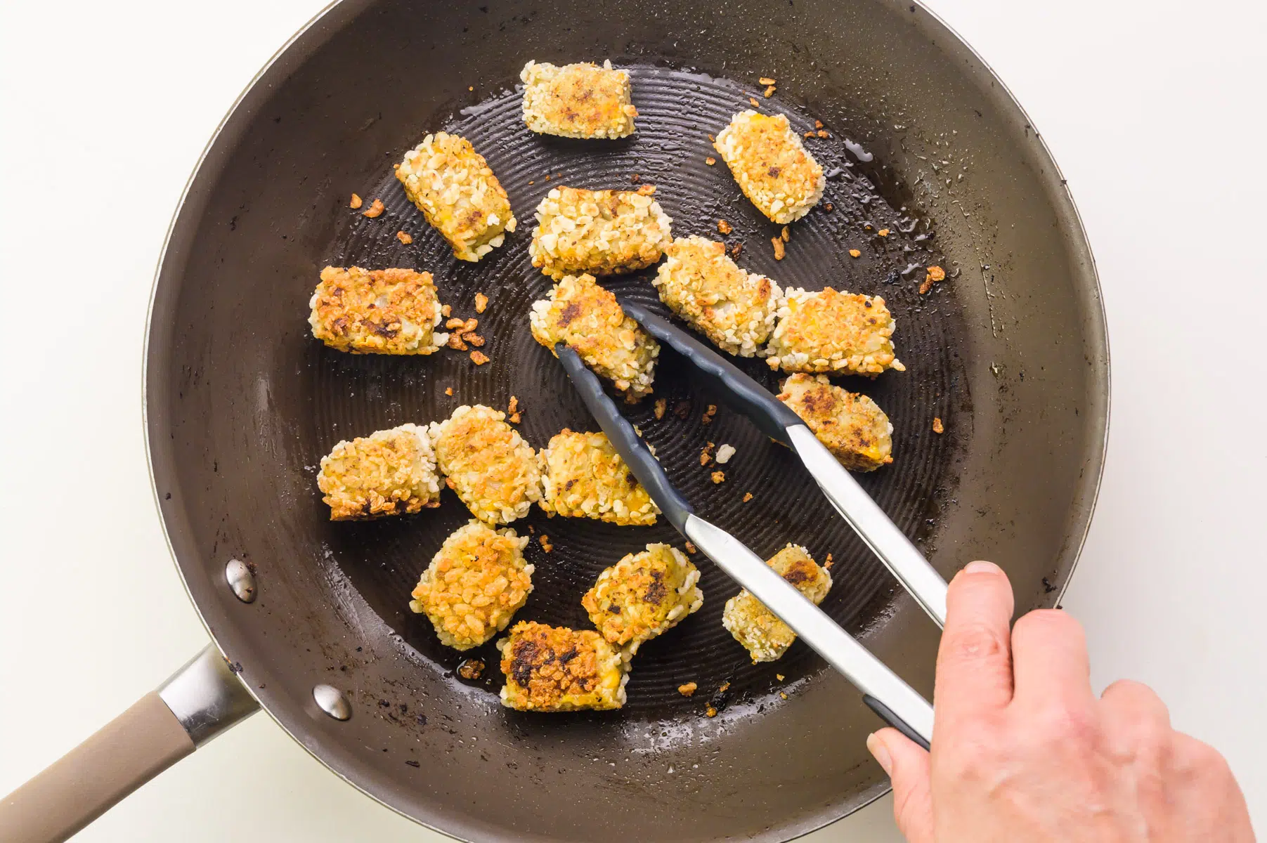 A hand uses tongs to turn cauliflower tater tots in a skillet.