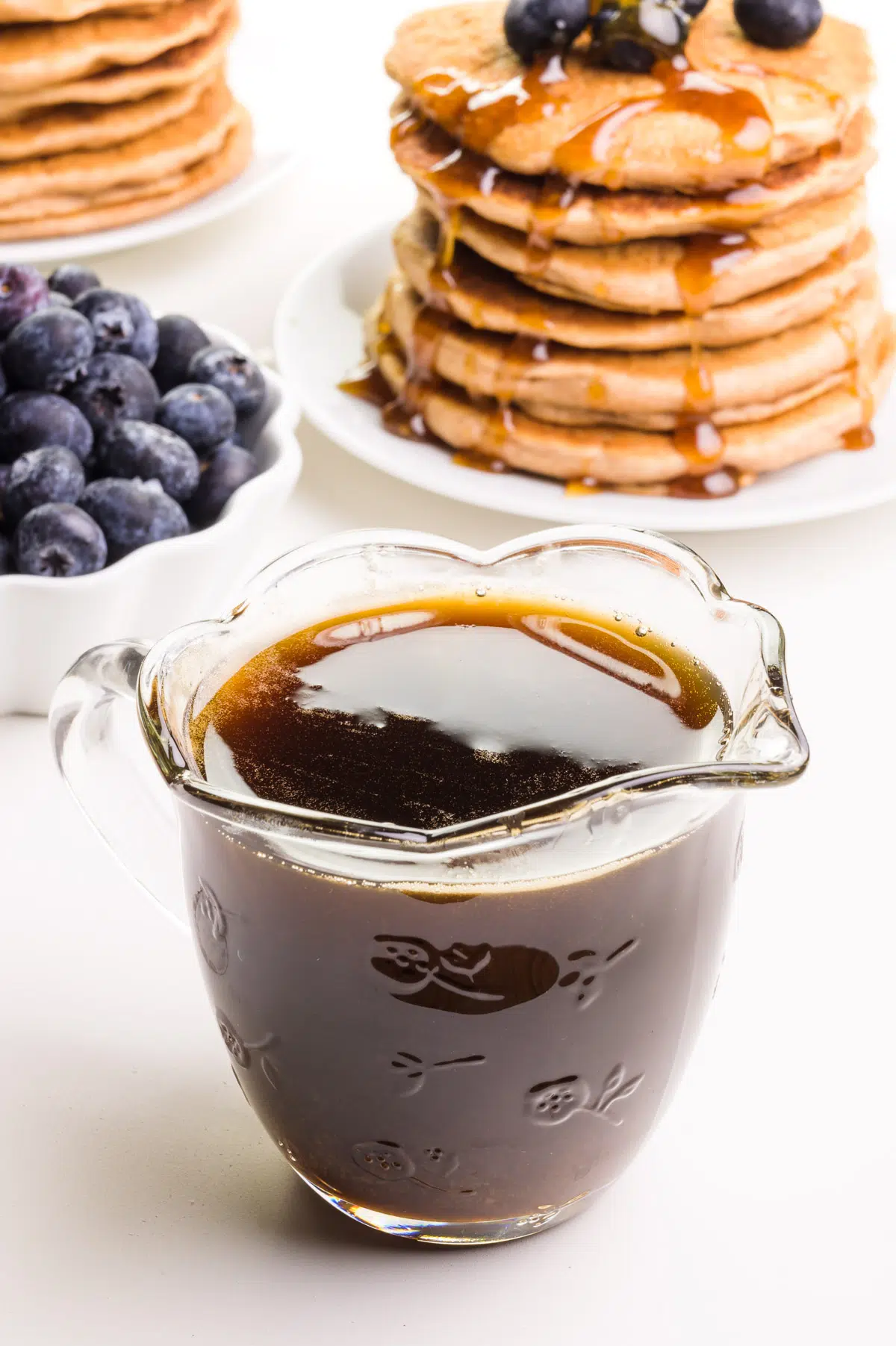 A glass pitcher of maple syrup substitute sits in front of a bowl of fresh blueberries and plates of pancakes.