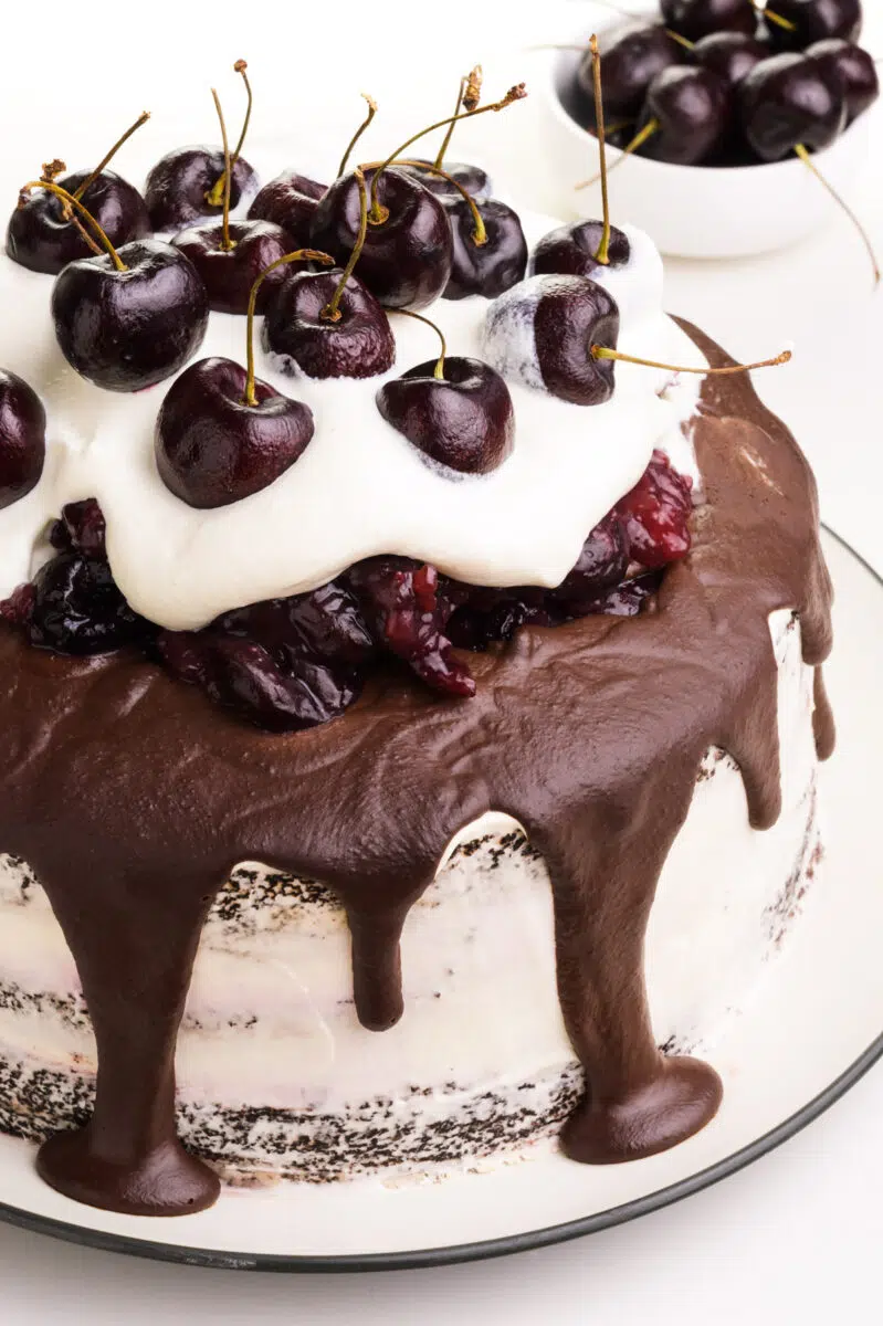 A vegan Black Forest cake has fresh cherries on top. There's a bowl of cherry sauce in the background.