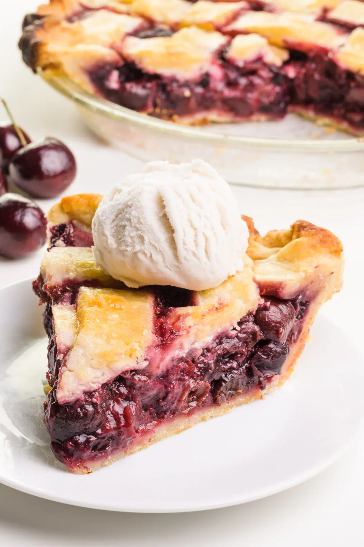 A slice of vegan cherry pie sits on a plate with a scoop of ice cream on top. There is a bowl of cherries and the rest of the pie in the background.