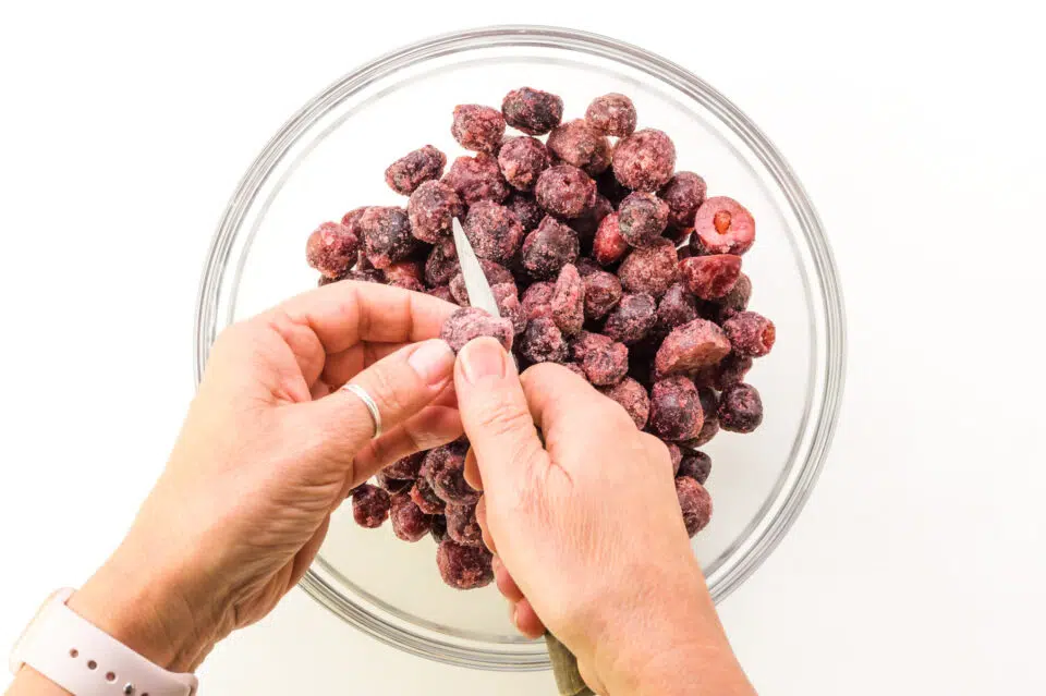 Two hands are cutting cherries in half, over a bowl full of frozen cherries.
