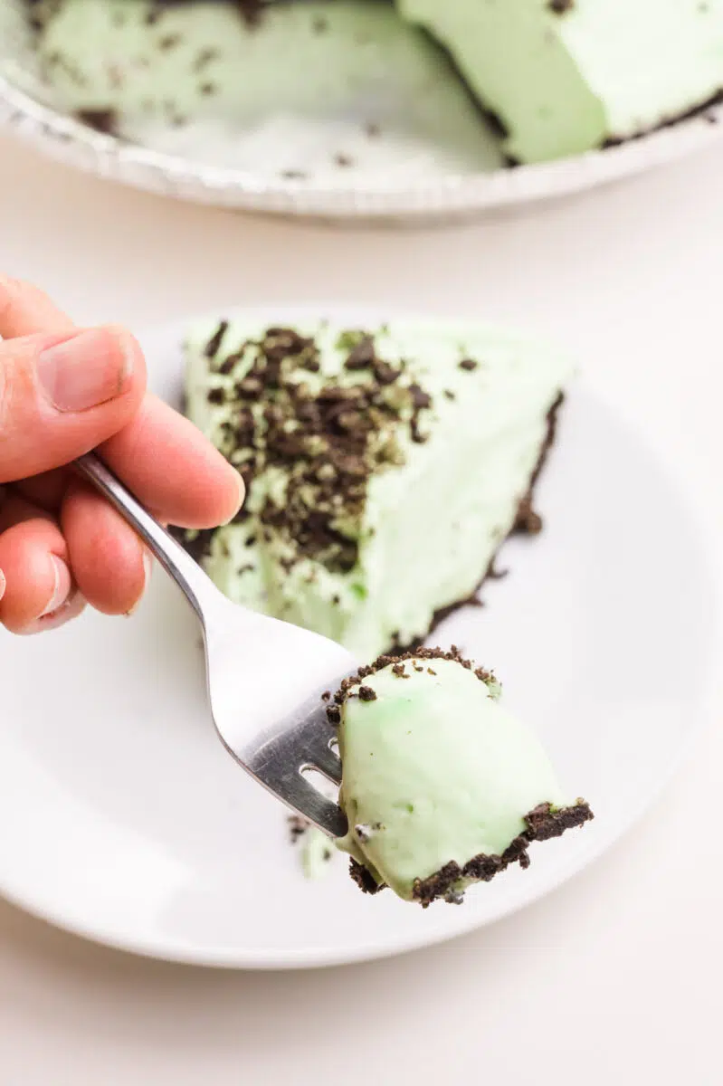 A hand holds a forkful of green dairy-free grasshopper pie hovering above the rest of the slice. The rest of the pie is in the background.