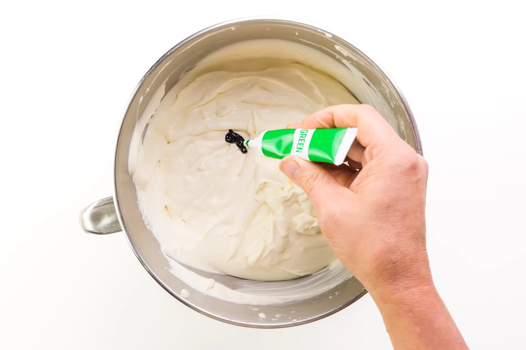 A hand holds a tube of green food coloring, adding drops to a bowl of whipped cream.
