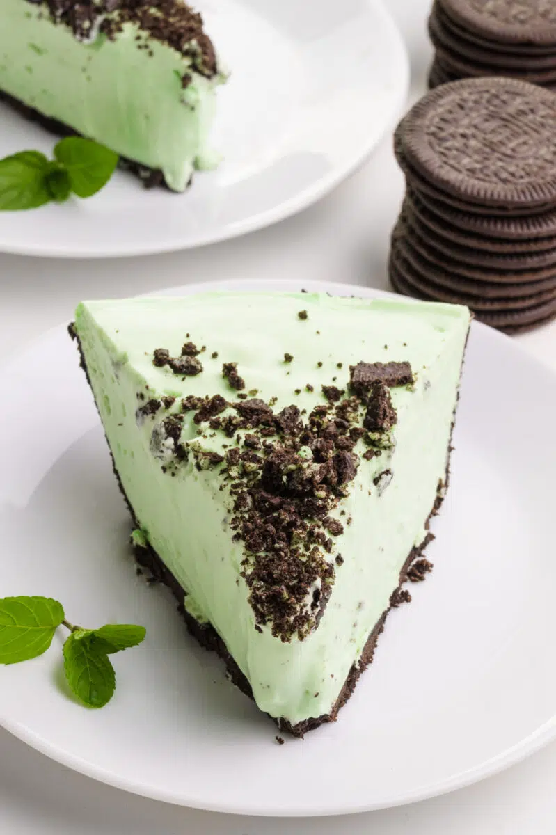 A slice of healthy grasshopper pie sits on a plate. There's another slice on a plate and some chocolate sandwich cookies behind it.
