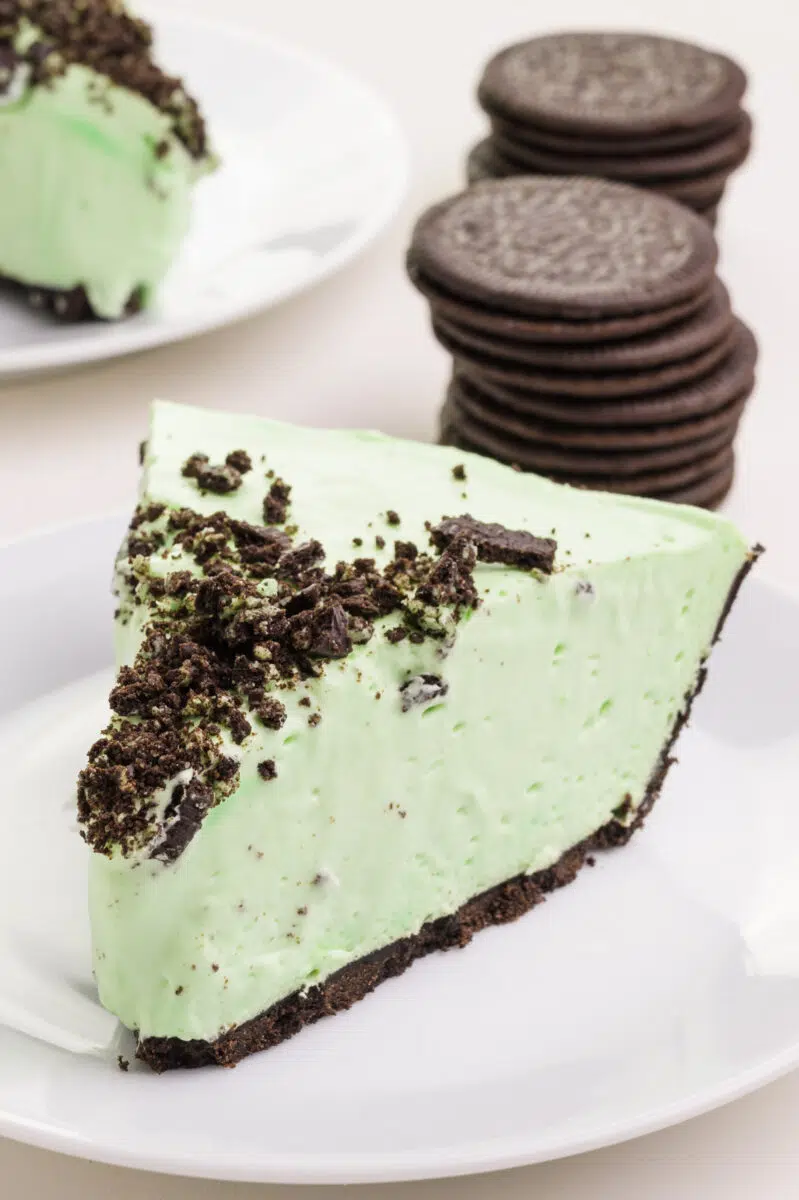A slice of vegan grasshopper pie sits in front of stacks of sandwich cookies and another slice of pie.