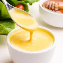 A spoon with vegan honey mustard dressing hovers over a bowl of it. There is a salad and a bowl with honey in the background.
