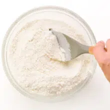 A hand holds a spatula, stirring together flour ingredients in a bowl.