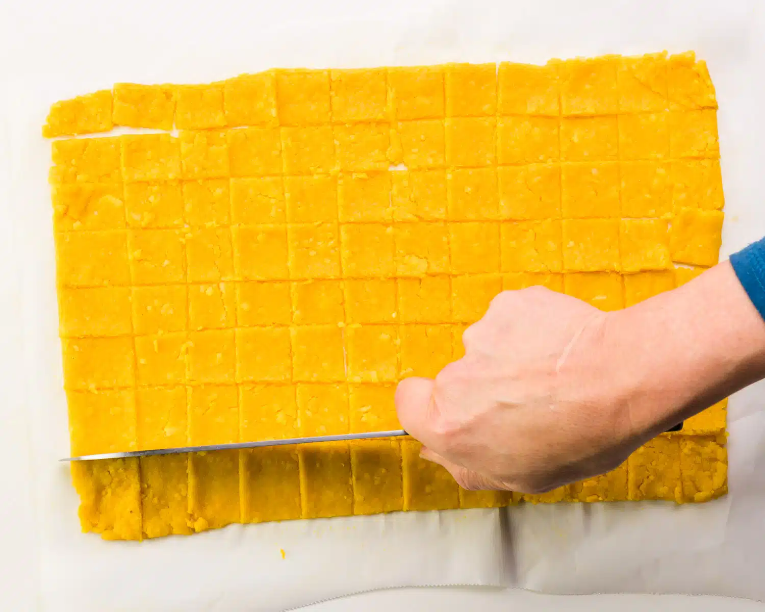 A hand holds a knife, cutting a thin orange dough into squares.