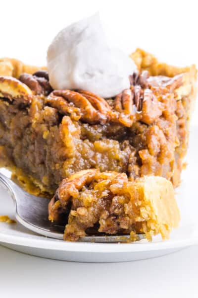 A closeup of a slice of vegan pecan pie on a plate. A bite is sitting on a fork in front of the plate.
