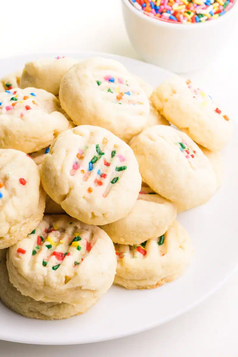 A plate of vanilla cookies sits in front of a bowl of sprinkles.