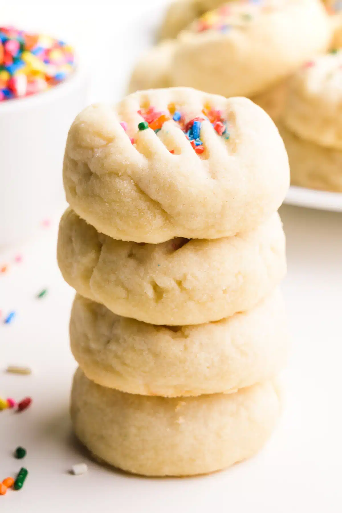 A stack of vegan shortbread cookies sits in a front of a bowl of sprinkles and a plate with more cookies.