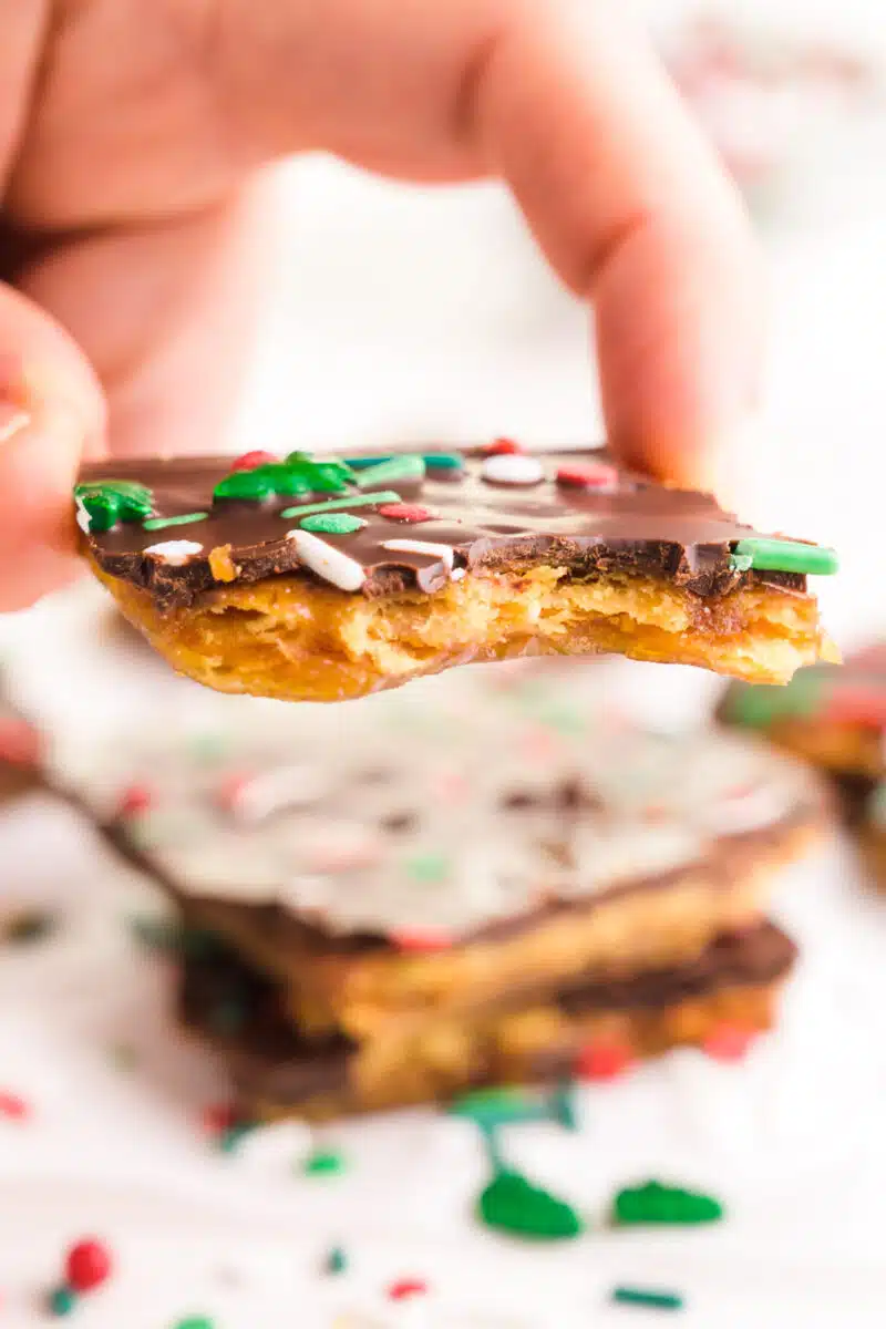 A hand holds a piece of Christmas crack. It hovers over a table with another slice and sprinkles all around.