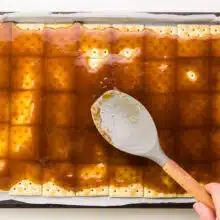 A hand holds a spatula, spreading caramel over crackers.