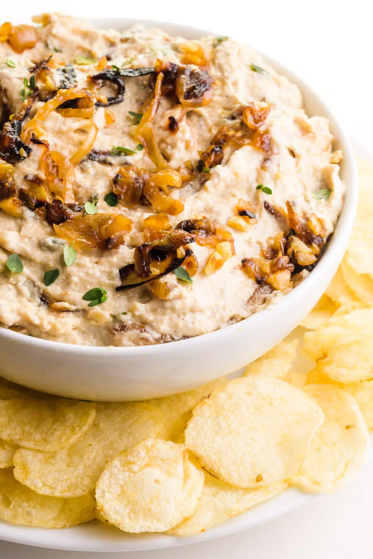 A bowl of vegan French onion dip sits on a plate with potato chips.