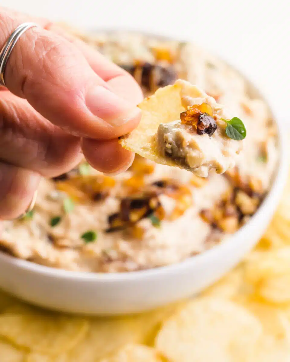 A hand holds a chip with dip on it. It hovers over a bowl with more creamy dip and potato chips.