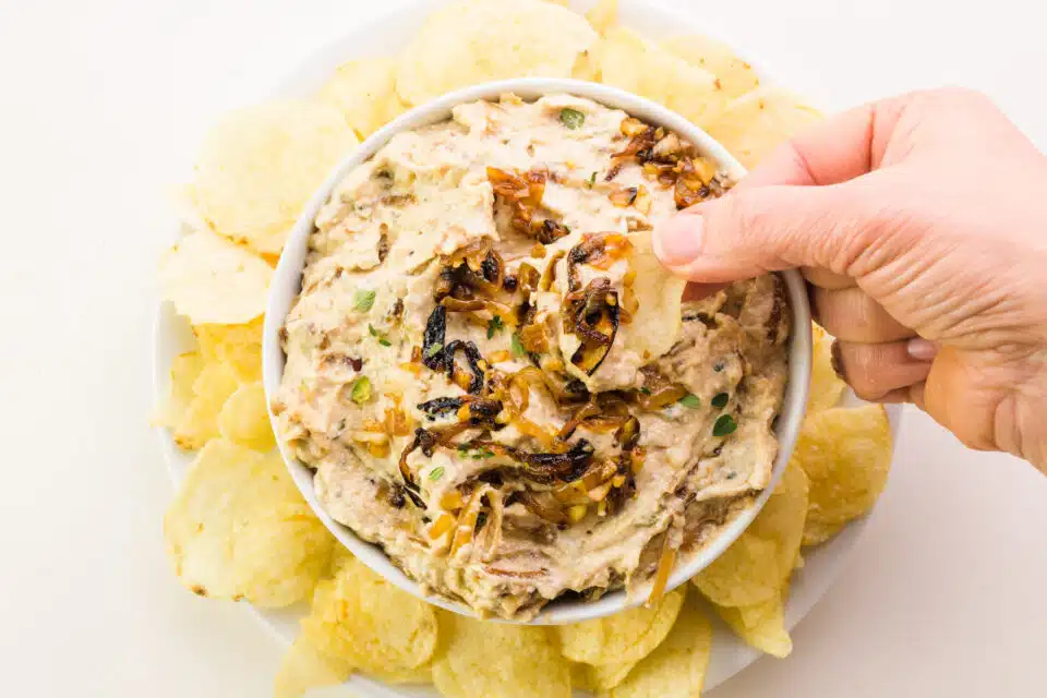 A hand holds a potato chip with dairy-free French onion dip. It hovers over a bowl of the dip surrounded by potato chips.