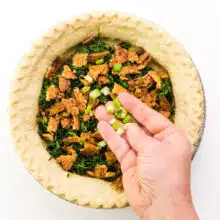 A hand spreads toppings over a pre-baked pie crust.