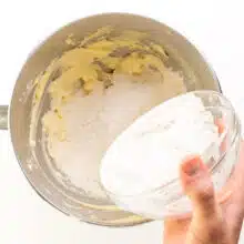 A hand holds a bowl, pouring flour into a mixing bowl with whipped butter.