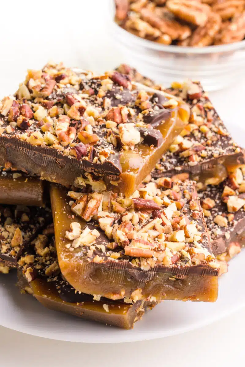 Chunks of homemade vegan toffee sits on a plate in front of a bowl of pecans.