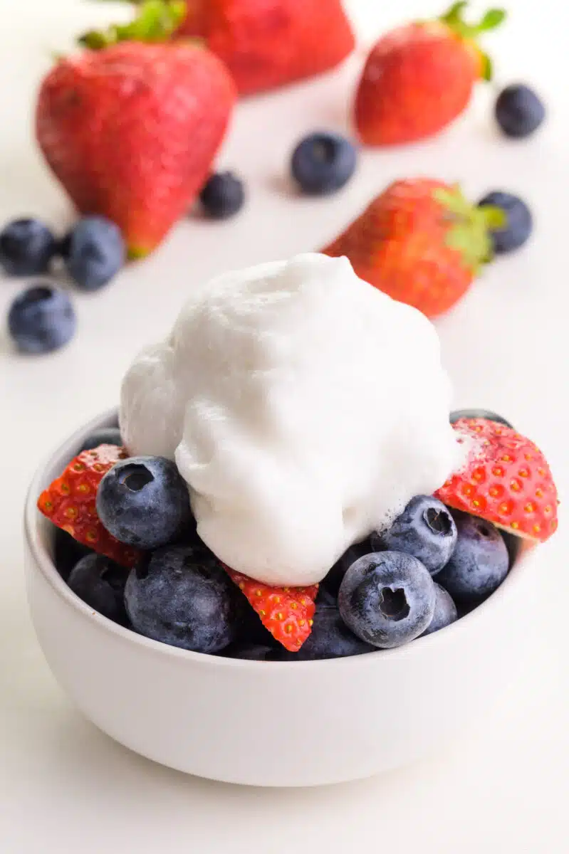 A bowl of berries is topped with whipped cream. There are more berries in the background.