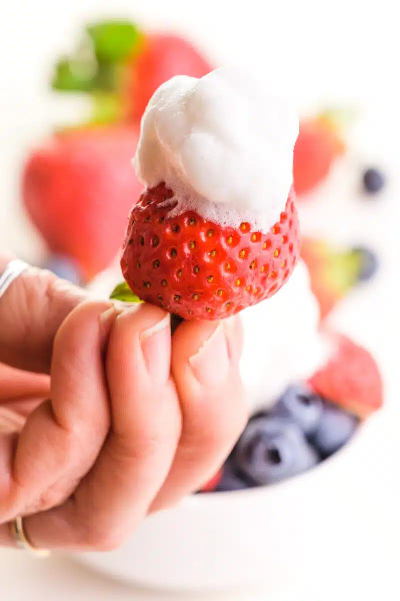 A hand holds a strawberry topped with whipped cream. There are more berries in the background.