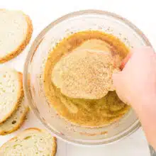 A hand places a slice of bread in a bowl, dunking it in a liquid mixture. There are more slices of bread beside the bowl.
