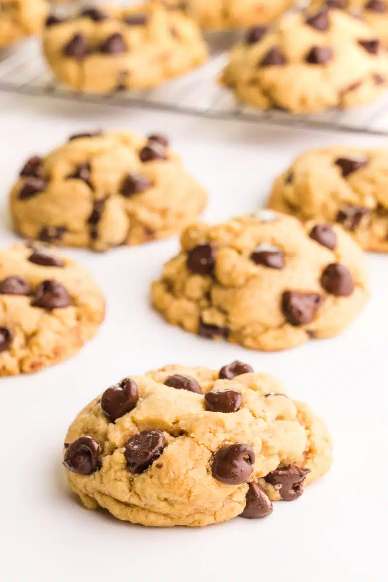 Several chocolate chip cookies sit on a table in front of a rack with more cookies.