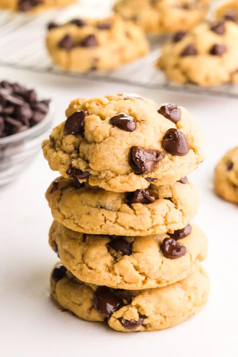 A stack of vegan peanut butter chocolate chip cookies sits beside a bowl of chocolate chips and more cookies in the background.