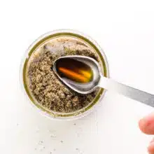 A hand holds a measuring spoon with vanilla, pouring it into a bowl with chia seeds.