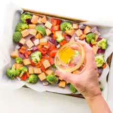 A hand pours olive oil over veggies in a sheet pan.