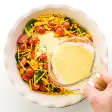 A hand pours Just Egg mixture over veggies and cheese in a pie pan.