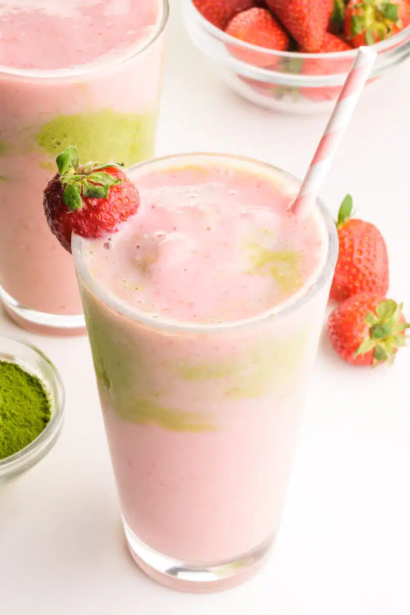 A closeup of a strawberry smoothie with matcha in it. There is another smoothie and fresh strawberries in the background.