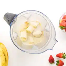 Looking down on a blender with ingredients like banana chunks. There are fresh strawberries and bananas around the blender.