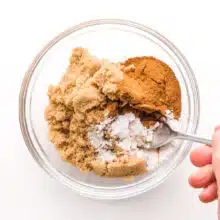 A hand holds a spoon, stirring cinnamon, brown sugar, and other ingredients in a bowl.
