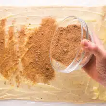 A hand holds a bowl with a cinnamon mixture, sprinkling it over rolled out dough.