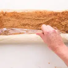 A hand holds plastic wrap, using it to roll a long sheet of dough sprinkled with cinnamon sugar.