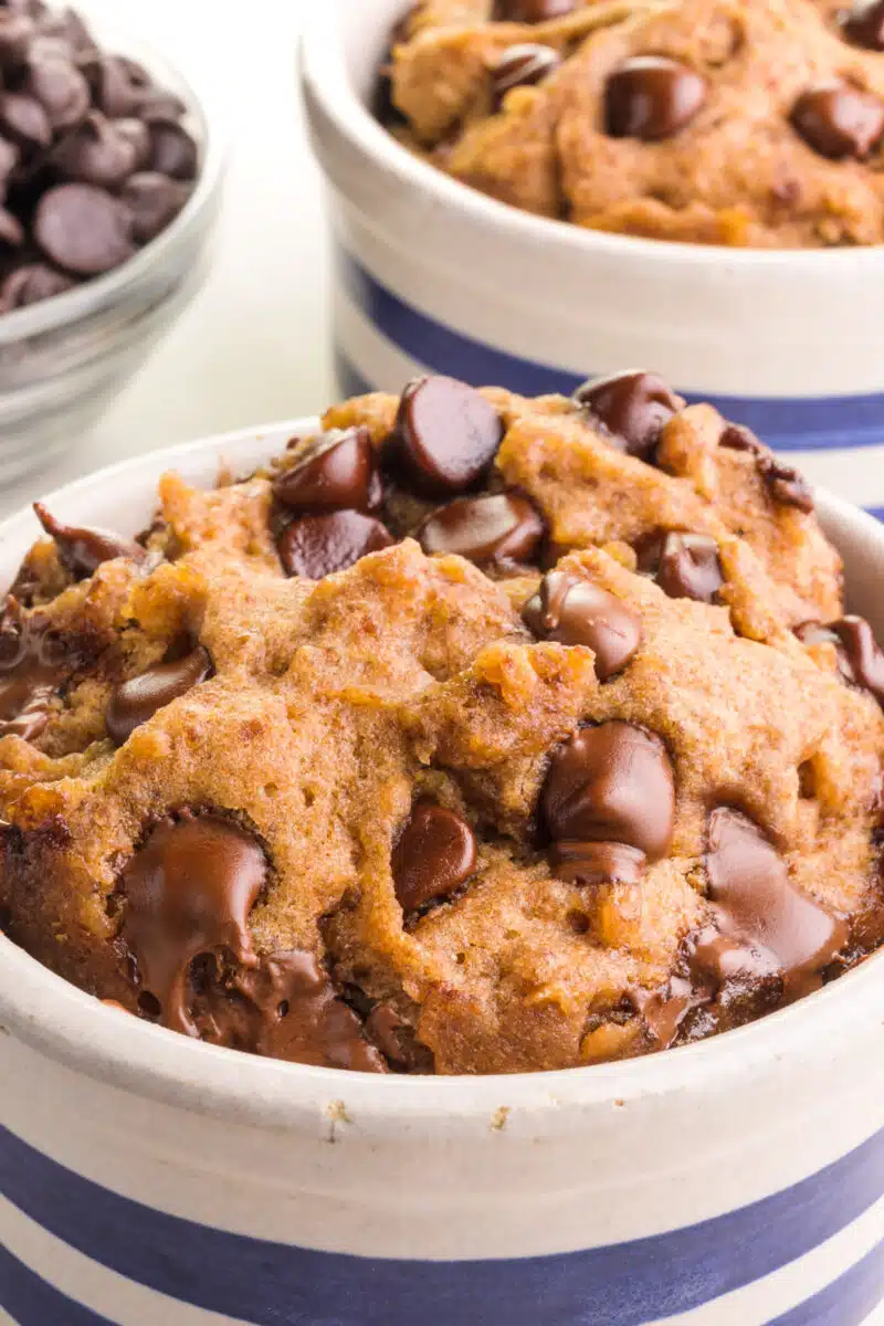 A closeup of a mug cookie with another one and a bowl of chocolate chips behind it.