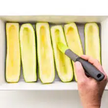 A hand holds a melon scooper, scooping flesh out of zucchinis in a baking dish.
