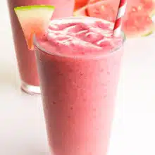 Two pink smoothies sit on a table. They have watermelon wedges and paper straws. There are watermelon wedges in the background.
