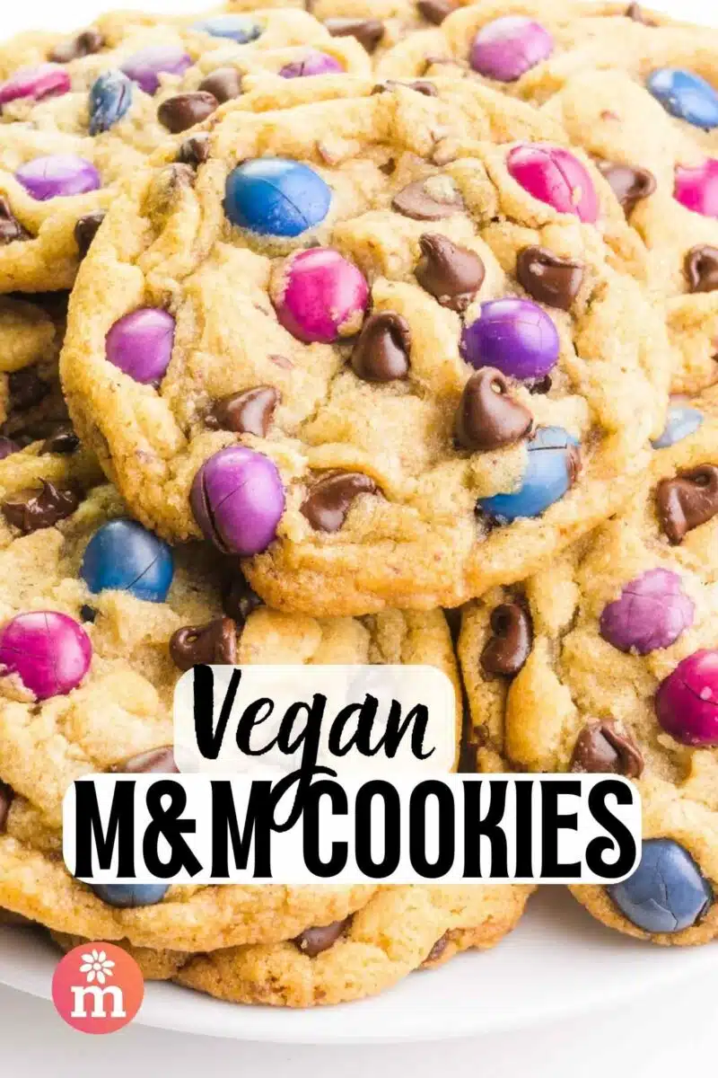 A plate of cookies has colorful candies and chocolate chips. The text reads, Vegan M&M Cookies.