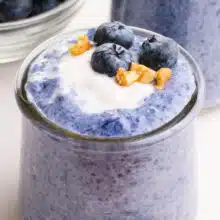 A closeup of blue chia pudding in a glass jar. It is topped with vegan yogurt, chopped walnuts, and fresh blueberries.